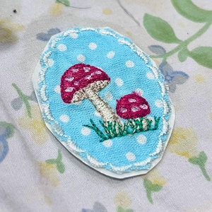 May include: An embroidered patch featuring two red mushrooms with white spots on a light blue background with white polka dots. The mushrooms are set on green grass, and the patch has a white scalloped border. The patch is an oval shape.