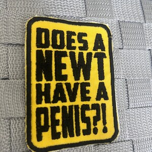 Peut inclure: Un écusson brodé rectangulaire avec un fond jaune vif et une bordure noire épaisse. L'écusson présente des lettres noires en gras de style bloc qui indiquent : DOES A NEWT HAVE A PENIS?! Le texte est centré et remplit toute la surface jaune.