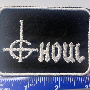 May include: A rectangular black embroidered patch with a white border, featuring a white cross-like symbol and the text "HOUL" in white stitching. A blue ruler is positioned at the bottom, showing the patch is approximately 3 inches wide.