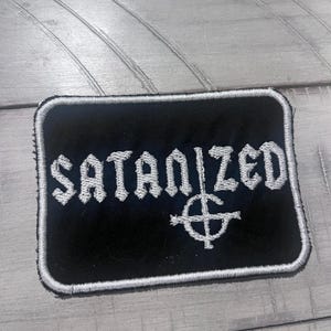 May include: A black rectangular patch with the word "SATANIZED" embroidered in white, along with a cross symbol. The patch has a white border and is designed to be sewn onto clothing or accessories.
