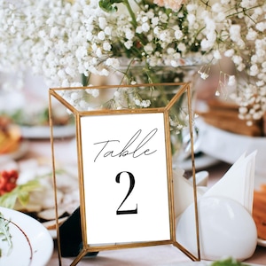 May include: A gold-framed table number card with the word "Table" and the number "2" in black script. The card is displayed on a table with white flowers in the background, suggesting a wedding or event setting.