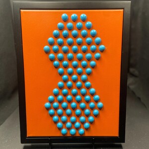 Könnte beinhalten: Abstrakte Wandkunst mit einem leuchtend orangefarbenen Hintergrund und einem rautenförmigen Muster aus türkisfarbenen Kugeln. Das Kunstwerk ist in einem schwarzen rechteckigen Rahmen gefasst und auf einer schwarzen Staffelei ausgestellt. Ca. 20 cm x 20 cm.