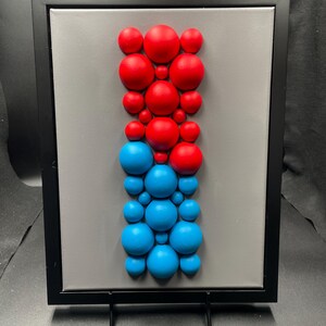 Può includere: Opera d'arte astratta con cornice nera, sfondo grigio e una composizione verticale di sfere rosse e blu. Le sfere variano in dimensioni, creando una composizione dinamica. L'opera è esposta su un cavalletto nero, adatta per l'arredamento.