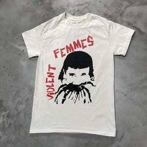 May include: Off-white t-shirt with a graphic design. The design features the words "FEMMES" and "VIOLENT" in red, with a black illustration of a person's face and hands. The shirt is made of a soft material.