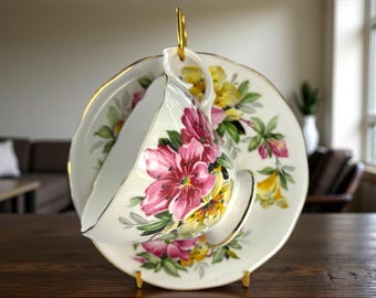 May include: A vintage teacup and saucer set with a floral design. The cup and saucer are white with pink, yellow, and green floral patterns and gold trim. The teacup is suspended on a gold-colored stand.
