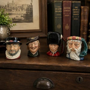 May include: Four ceramic character jugs arranged in a row. The first is a person in a black hat and white ruff. The second is a person in a brown hat. The third is a guard in a tall black bearskin hat and red uniform. The fourth is a bearded figure.