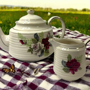 May include: A white ceramic teapot and creamer set with a floral rose design. The teapot has a lid and a curved spout. The creamer has a handle. Both pieces have a ribbed texture and gold trim. The set is on a checkered cloth.