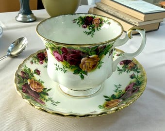 May include: White teacup and saucer set with a floral rose pattern in shades of red, yellow, and green. The cup and saucer have gold trim. The teacup is sitting on a white tablecloth.