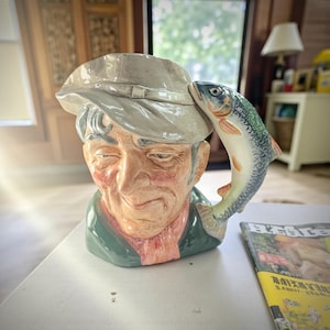 May include: A ceramic mug shaped like an elderly man wearing a hat, with a fish forming the handle. The mug features a detailed, hand-painted design with a green coat, pink shirt, and a gray hat. The fish is blue, white, and orange.