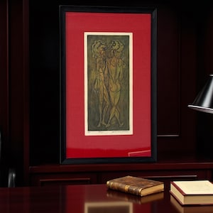 May include: Framed artwork showing a print of three figures with stylized bodies in shades of green and tan. The artwork is set against a red background within a black frame. The piece is displayed on a dark wood surface.
