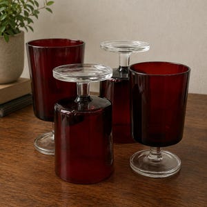 Op de afbeelding: Een set van vier dieprode glazen bekers met heldere glazen stelen en ronde voeten. Twee van de glazen staan rechtop, terwijl de andere twee ondersteboven op hun rand staan, waardoor de heldere glazen voeten en stelen zichtbaar zijn.