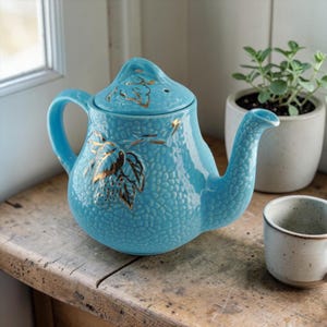 Könnte beinhalten: Türkisfarbene Keramik-Teekanne mit strukturierter Oberfläche und goldenem Blattdesign. Die Teekanne hat einen gebogenen Ausguss, einen Henkel und einen Deckel. Eine kleine Teetasse steht daneben auf einer Holzoberfläche.
