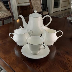 Puede incluir: Un juego de té de porcelana blanca con tetera, jarra de crema, azucarero, taza y platillo. Cada pieza tiene un borde plateado y un patrón texturizado. El juego está dispuesto sobre una mesa de madera oscura.