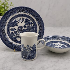 Op de afbeelding: Een witte keramische mok, bord en kom set met een blauw Willow patroon. Het bord en de kom hebben een geschulpte rand. De mok, het bord en de kom zijn gerangschikt op een lichtgekleurd oppervlak. Een groene plant in een witte pot staat op de achtergrond.