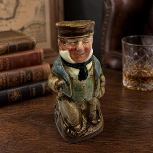 May include: A ceramic character jug shaped like a person wearing a brown cap, a blue jacket, and a black neckerchief. The figure has a light-coloured collar, a ruddy complexion, and is depicted standing with one hand resting on a coiled rope base.