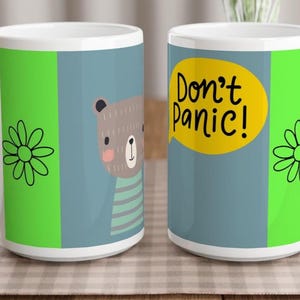 Puede incluir: Dos tazas de cerámica blanca con un diseño gráfico sobre fondo azul, una ilustración de un oso pardo y un bocadillo amarillo con el texto negro Don't Panic!. Las tazas también muestran paneles laterales de color verde brillante con flores negras.