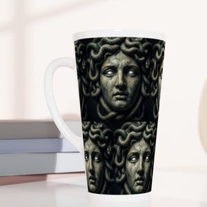 Könnte beinhalten: Ein hoher weißer Keramik-Latte-Becher mit einem sich wiederholenden dunklen Grafikdruck des Medusa-Kopfes mit steinartiger, rissiger Haut, leuchtend weißen Augen und Schlangen als Haaren. Der Becher hat einen glatten weißen Henkel.
