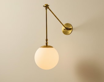 May include: A brass-toned wall sconce with a white globe-shaped light fixture. The sconce features an adjustable arm and a round wall plate. The light fixture is a sphere, and the overall design is modern and minimalist.