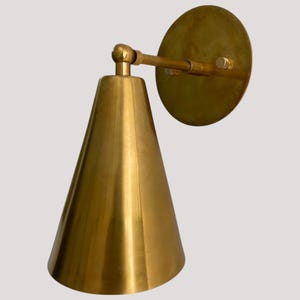 May include: A brass wall sconce with a conical shade and a circular backplate. The sconce has a warm, golden hue and a minimalist design. The shade is approximately 6 inches in diameter at its base. The fixture is mounted on a wall.
