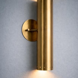 May include: A cylindrical wall sconce made of brushed brass with a circular mounting plate. The fixture features a series of small, evenly spaced perforations along the top and bottom edges, emitting a warm, focused light from both ends.