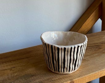May include: A small, off-white ceramic bowl with a black vertical stripe pattern. The bowl has an irregular, slightly wavy rim and sits on a wooden shelf. The interior of the bowl is smooth and glossy.