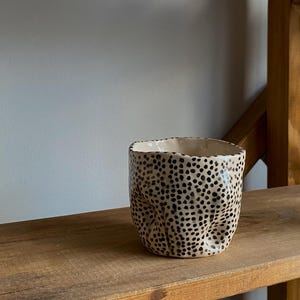 Könnte beinhalten: Eine beige Keramiktasse mit strukturierter Oberfläche und schwarzen Tupfen. Die Tasse hat eine einzigartige, leicht zerknitterte Form und steht auf einem Holzregal. Es ist ein dekorativer Gegenstand.