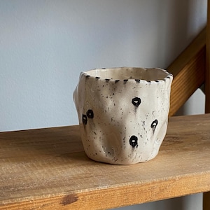 Pode incluir: Uma caneca de cerâmica artesanal com uma superfície texturizada em branco sujo e formato irregular. A caneca apresenta pontos pretos espalhados e uma borda escura. Está sobre uma prateleira de madeira, mostrando seu design único e orgânico.