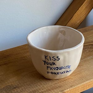 May include: Cream-coloured ceramic bowl with the handwritten text "KISS YOUR FAVOURITE FOREHEAD" in blue. The bowl is displayed on a wooden shelf.