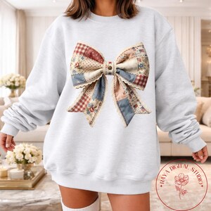 Puede incluir: Sudadera gris claro con un gran diseño de lazo de patchwork. El lazo incorpora varios patrones, incluyendo telas florales, a cuadros y de lunares en tonos de rojo, azul y crema. El lazo está adornado con encaje y tiene un botón en el centro.