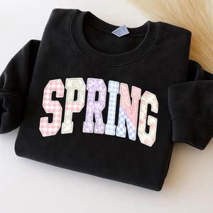 Può includere: Felpa nera con la scritta "SPRING" in un design patchwork sul davanti. Le lettere sono in colori pastello e presentano vari motivi, tra cui quadretti vichy e stampe floreali. La felpa ha un girocollo e maniche lunghe.
