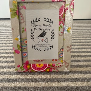 May include: A rectangular photo frame with a floral pattern in pink, green, and yellow on a beige background. The frame has the text "From Paola With Love" and "Quirky Paper Goods and more".