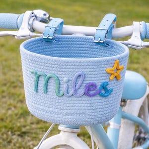 Op de afbeelding: Een lichtblauwe fietsmand met de naam "Miles" in kleurrijke garenletters en een gele zeester. De mand is bevestigd aan het stuur van een lichtblauwe fiets. De mand is gemaakt van geweven materiaal.