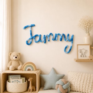 May include: A wall-mounted name sign spelling the name Tammy in cursive script, crafted from a thick, knitted yarn in alternating shades of light and dark blue. The letters are connected in a single continuous line, creating a soft, textured appearance.