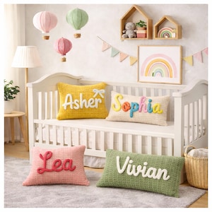 May include: A nursery scene with a white crib, featuring personalized name pillows in yellow, cream, pink, and green. Decorative hot air balloons hang from the ceiling. A rainbow art print and wooden shelves with decor complete the room.