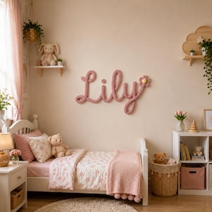 May include: A wall-mounted name sign spelling Lily in dusty pink knitted yarn, featuring a small crocheted flower accent. The script-style letters are connected in a continuous loop, creating a soft, textured decorative piece for a bedroom wall.