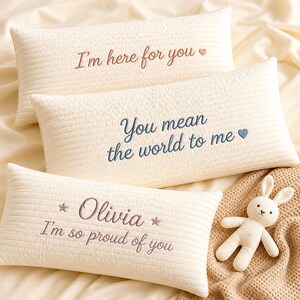 Peut inclure: Trois coussins rectangulaires matelassés de couleur crème avec du texte brodé. Le coussin du haut porte l'inscription "I'm here for you" avec un cœur. Celui du milieu "You mean the world to me" avec un cœur. Celui du bas "Olivia I'm so proud of you" avec des étoiles.