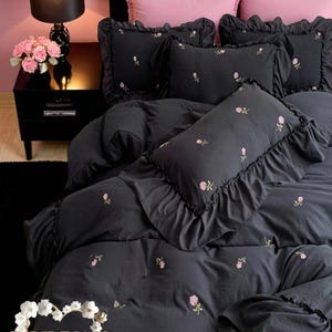 May include: Dark gray bedding set with pink floral embroidery. The set includes a duvet cover, ruffled pillowcases, and pink accent pillows. A black nightstand with a lamp and flowers is visible in the background.