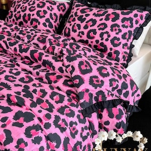 May include: A pink leopard print bedding set with black accents. The set includes a comforter and two pillowcases. The pillowcases and comforter have a black ruffle trim. The bedding is on a black bed frame.