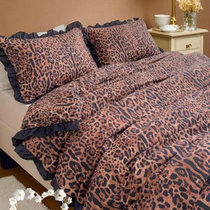 Könnte beinhalten: Bettwäsche-Set mit Leopardenmuster in Braun- und Schwarztönen. Das Set beinhaltet einen Bettbezug und zwei Kissenbezüge mit dunkelblauer Rüschenborte. Die Bettwäsche ist auf einem Bett mit weißer Matratze drapiert. Das Thema ist Animal Print.