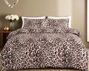 Puede incluir: Una cama con un edredón con estampado de leopardo y dos fundas de almohada a juego. La ropa de cama presenta un patrón de manchas marrones y negras sobre un fondo beige. La cama está hecha y lista para usar.