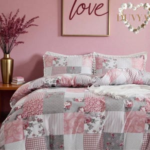 May include: A patchwork quilt-style bedding set in shades of pink, grey, and white. The design features floral patterns, stripes, and polka dots. The headboard has two matching pillowcases. A gold vase with pink flowers sits on a wooden table.