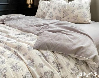 May include: A bed is made with a floral patterned duvet cover and pillowcases. The bedding features a cream background with gray rose illustrations. A gray blanket is folded over the duvet. A gold vase with flowers sits on a black table.