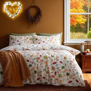 Op de afbeelding: Witte beddengoedset met een patroon van herfstbladeren en pompoenen. De set bevat een dekbedovertrek, kussenslopen en een bruine deken. De kamer heeft een herfstthema met uitzicht op herfstbomen door een raam.