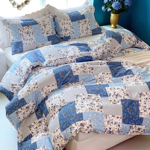 Pode incluir: Um conjunto de colcha e almofadas de patchwork em tons de azul, branco e cinza. A roupa de cama apresenta padrões florais e paisley, criando uma estética acolhedora. A palavra "DUVVAY" é visível no canto superior esquerdo.
