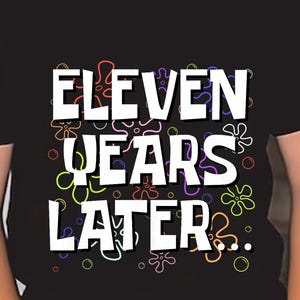Op de afbeelding: Zwart t-shirt met de tekst "ELEVEN YEARS LATER" in het wit, met kleurrijke bloemaccenten. Het shirt wordt gedragen door een kind met een zwarte zonnebril.