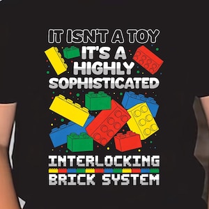 May include: A black t-shirt featuring a colorful graphic of interlocking toy bricks. The shirt has the text "IT ISN'T A TOY IT'S A HIGHLY SOPHISTICATED INTERLOCKING BRICK SYSTEM". The shirt is worn by a child wearing black sunglasses.