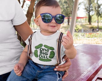 May include: A baby wearing blue sunglasses, a white t-shirt with a "One Big Dill" graphic, and suspenders. The baby is smiling and being held by an adult. The background is a park setting with trees and a red picnic table.