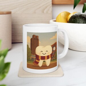 May include: White ceramic mug featuring a cartoon tooth wearing a scarf with a cityscape background. The tooth has a smiling face and is wearing a red, brown, and gold scarf. The background includes buildings, trees, and a blue sky.