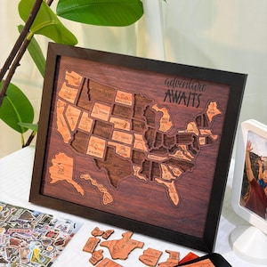 May include: A wooden map of the United States with individual state cutouts, framed in black. The words "adventure awaits" are written on the map. Additional wooden state cutouts and a black tin with the same phrase are also present.
