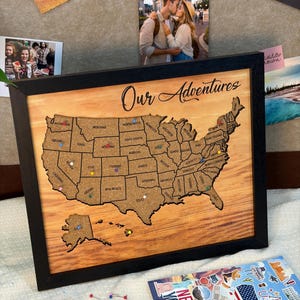May include: A framed wooden map of the United States with a corkboard center. The map is labeled "Our Adventures" and has colorful push pins marking locations. Photos and stickers are displayed around the map.